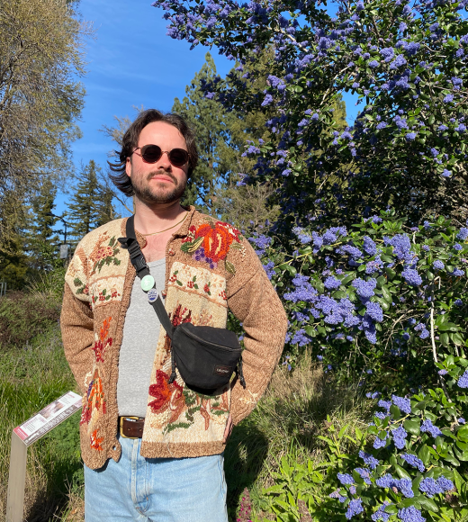 A man wearing a tan floral jacket stands in front of a tree