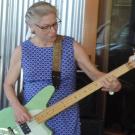 A woman with glasses wearing a blue dress with white dots playing an electric guitar.